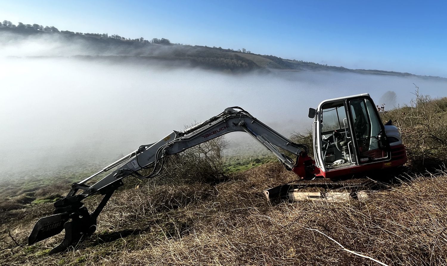 Tracked excavator with tree shear clearing scrub on a downland hillside above the mist